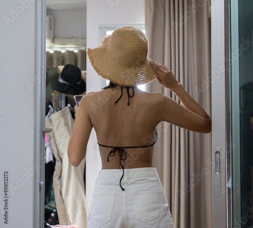 An Asian woman wearing white jeans is trying out a bikini and straw hat in her bedroom for a beach holiday.