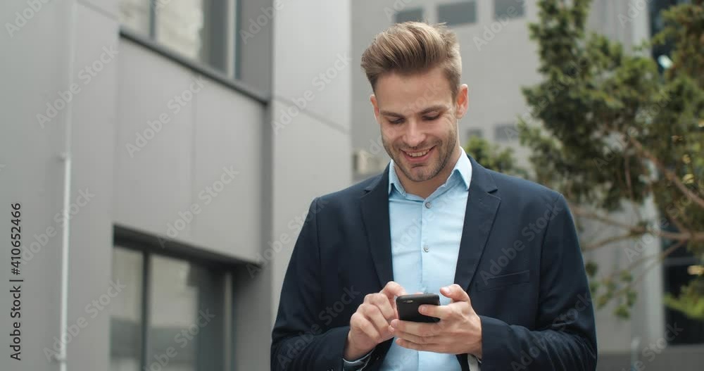 Handsome Man in Suit is smiling Charmingly while browsing Internet on his Phone. Successful Businessman is texting Messages while walking outside outdoor in modern Business District. People. Apps. Handsome Man in Suit is smiling Charmingly while browsing Internet on his Phone. Successful Businessman is texting Messages while walking outside outdoor in modern Business District. People. Apps.