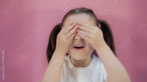 Little dark haired girl with two ponytails in white T-shirt plays hide and seek shows different emotions, makes peekaboo covering eyes with hands. Pink background. Concept of happy childhood lifestyle photo