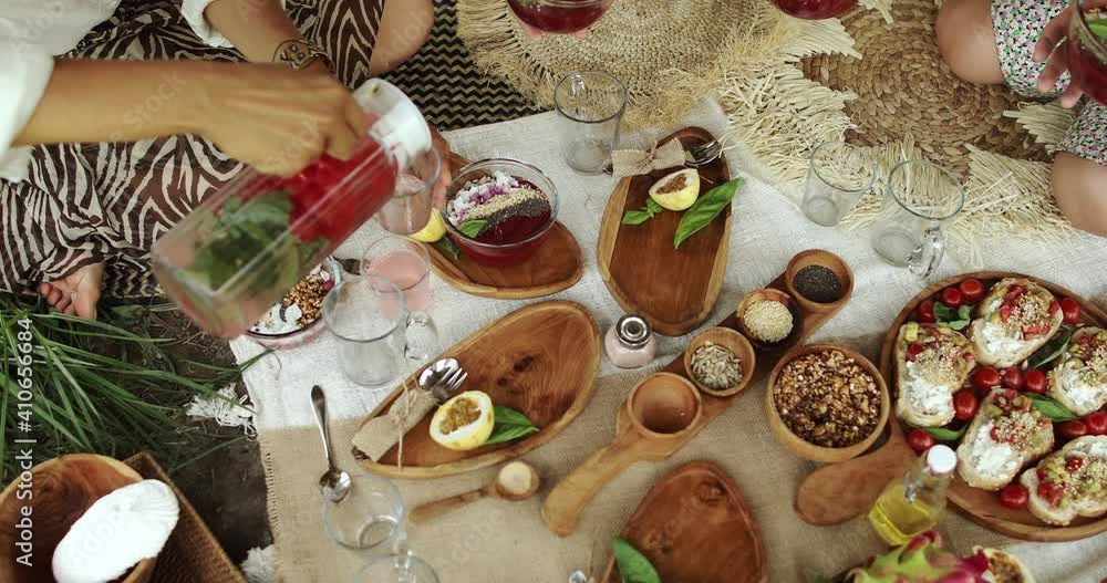 A group of girls sitting on a blanket with a plate of food at picnic taking the plates with smoothie and pouring the drink A group of girls sitting on a blanket with a plate of food at picnic taking the plates with smoothie and pouring the drink