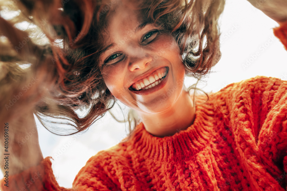 Bottom view of a candid beautiful young woman wearing a knitted orange sweater smiling broadly and looking directly to the camera outdoors. The pretty female has joyful expression, resting in the park - obrazy, fototapety, plakaty 
