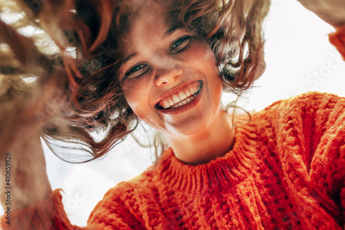 Bottom view of a candid beautiful young woman wearing a knitted orange sweater smiling broadly and looking directly to the camera outdoors. The pretty female has joyful expression, resting in the park photo