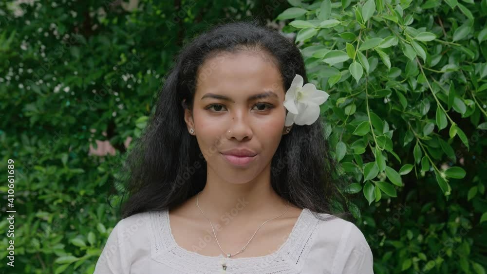 Portrait of a young beautiful Thai woman with a flower in her hair standing in a garden and looking straight into the camera on a sunny day in slow motion Portrait of a young beautiful Thai woman with a flower in her hair standing in a garden and looking straight into the camera on a sunny day in slow motion