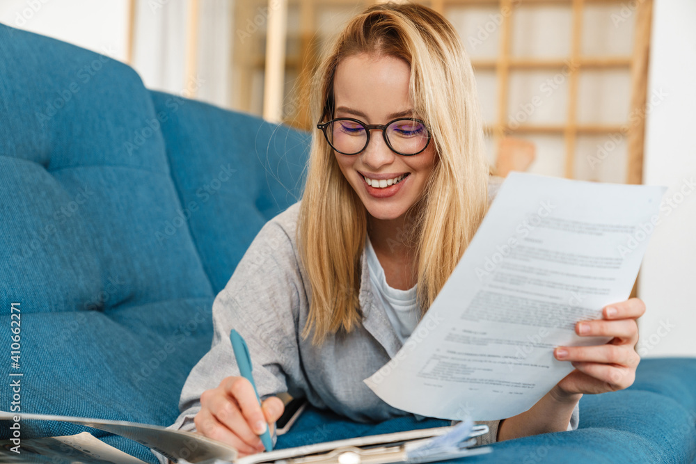 Smiling blonde woman in eyeglasses doing homework while lying on couch Smiling blonde woman in eyeglasses doing homework while lying on couch