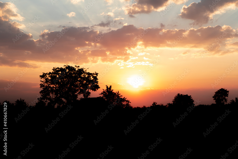 Silhouette of rural scenery with sunset in the mountains, red and yellow sky behind and trees with old cottage in the forefront