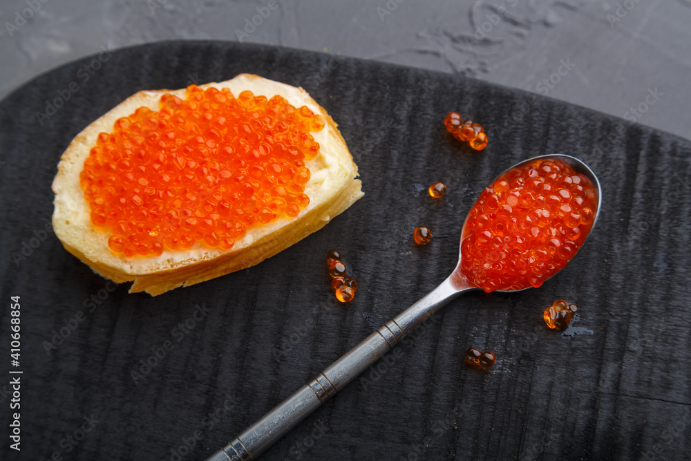 One toast with butter and red caviar next to a spoon with caviar on a wooden board on a concrete background. One toast with butter and red caviar next to a spoon with caviar on a wooden board on a concrete background.