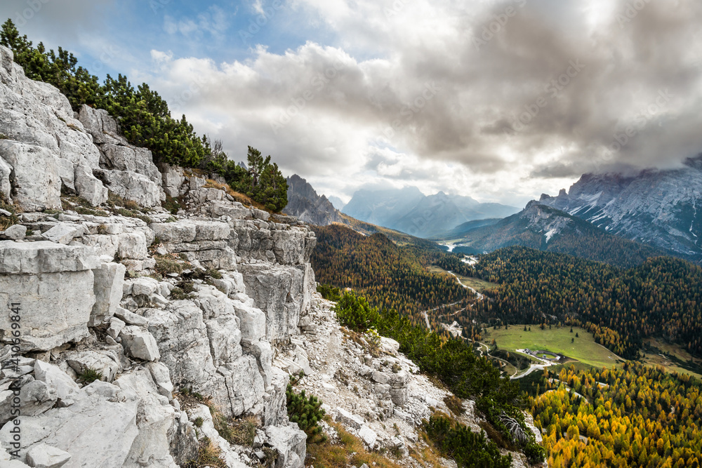 Wide angle view of a typical alpine landscape, with a rocky slope on the left and a valley covered by pine trees on the right, under puffy clouds