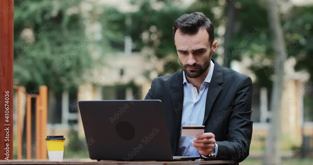 A man in a suit with a serious face enters numbers from a bank card while making a purchase on the Internet, sitting on the summer terrace of a cafe at a laptop. A man in a suit with a serious face enters numbers from a bank card while making a purchase on the Internet, sitting on the summer terrace of a cafe at a laptop.