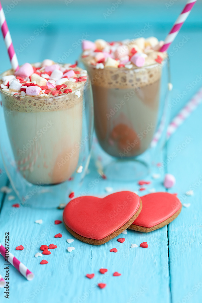Two glasses of hot coffee or cappuccino with marshmallows and heart-shaped cookies on a blue background. Two glasses of hot coffee or cappuccino with marshmallows and heart-shaped cookies on a blue background.