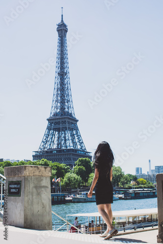 A girl in a black dress with long hair walking towards the river cruise Seine river and Eiffel Tower in Paris on a sunny day. © Alfred