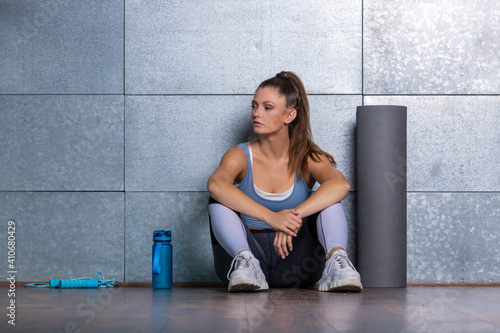 White ethnic fitness woman sitting on the floor arms rest on her spread legs looking to one site with pony tail mat bottle and rope on the floor beside her in front of a metal background, trainer tire