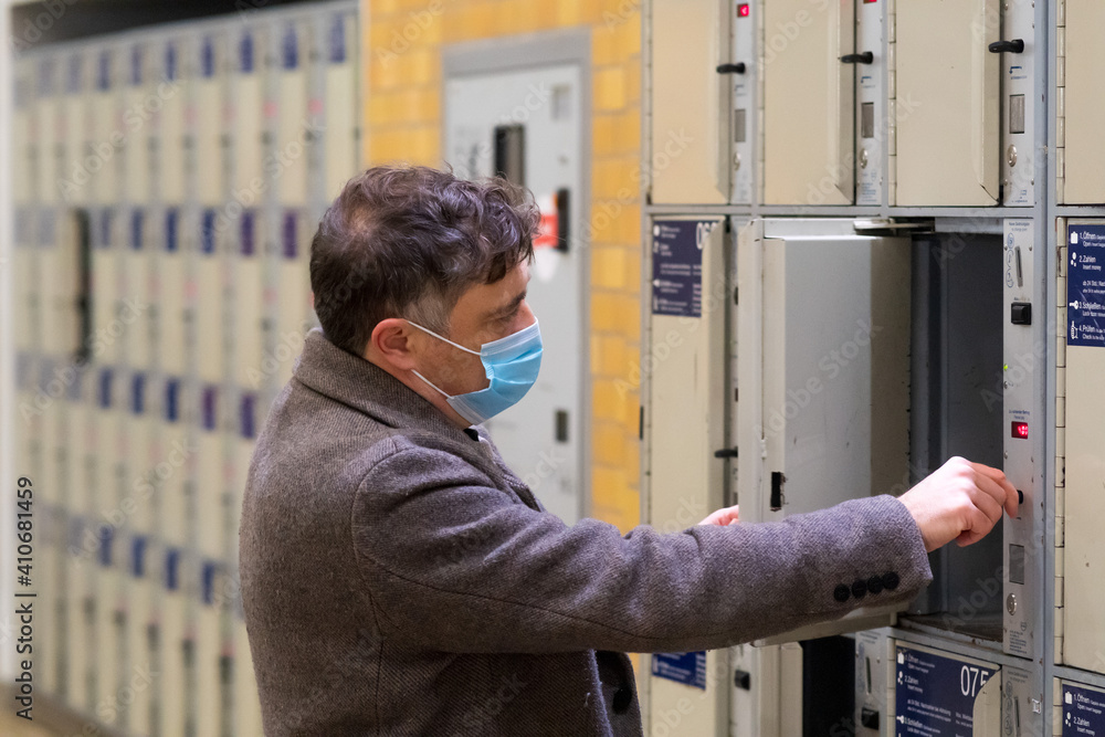 Male traveler wearing a protective surgical mask opening or closing a train station locker cabinet Male traveler wearing a protective surgical mask opening or closing a train station locker cabinet