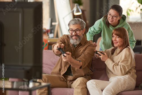 Two parents playing video game together sitting on sofa with their daughter standing in the background and watching for the game