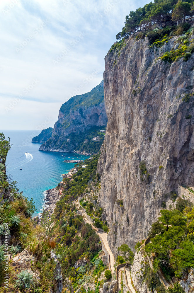 A magnificent landscape from Giardini di Augusto in Capri, Italy. Mountains, rocks, little road and the boat passing by in the background, followed by the clouds and sky.