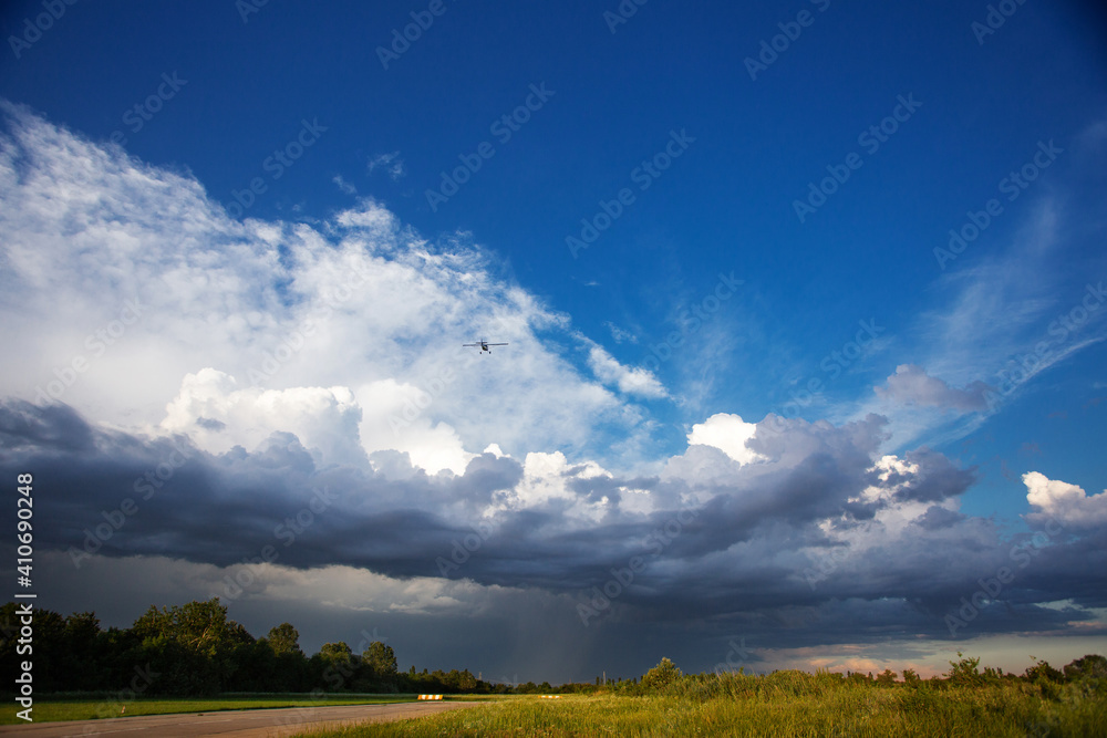 Small private plane climbs through the clouds. Aircraft is flying high above the beautiful landscape. Green field and runway in the sunset light after the thunderstorm Small private plane climbs through the clouds. Aircraft is flying high above the beautiful landscape. Green field and runway in the sunset light after the thunderstorm