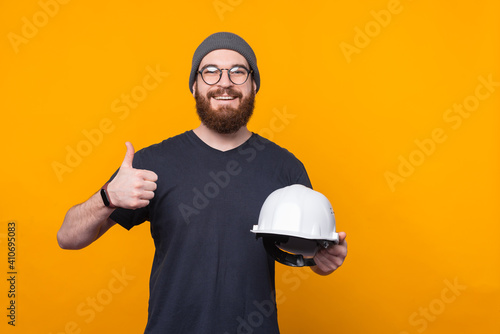 Photo of cheerful bearded man showing thumb up and holding helmet for construction.