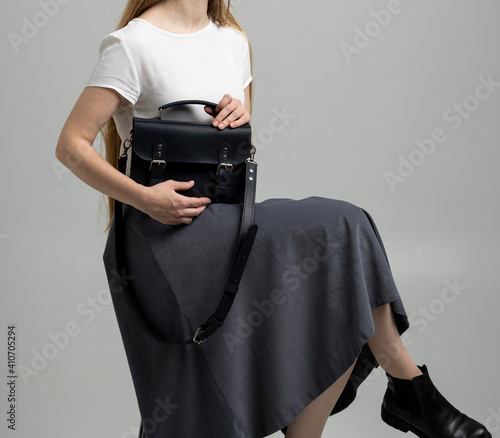Small black leather bag in a woman's hand on a white background. Shoulder handbag. Woman in a white shirt and grey skirt and with a black handbag. Style, retro, fashion, vintage and elegance.