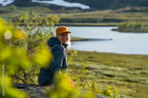 Fotografie Caucasian outdoor active young adult woman sitting on Rock With outdoor hiking cloths jacket and cap smiling and looking at camera with montain and lake background in Padjelanta national park, Sweden