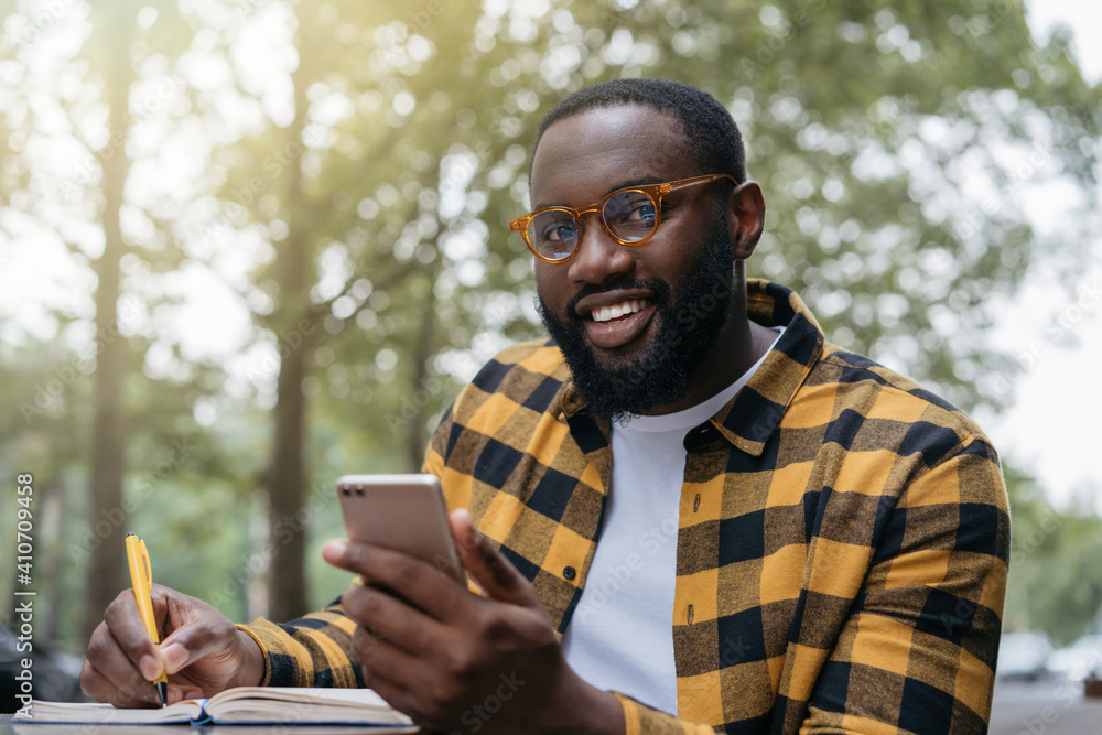 Smiling student studying, learning language, education concept. Confident African man using mobile phone, taking notes, working freelance project online Smiling student studying, learning language, education concept. Confident African man using mobile phone, taking notes, working freelance project online