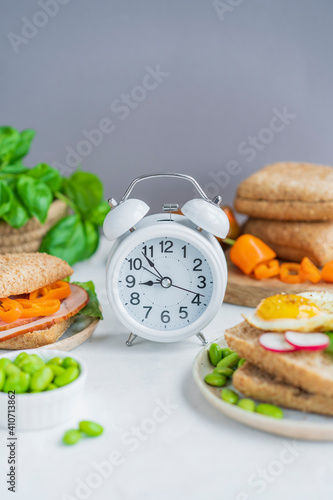 Alarm clock, healthy sandwiches with whole grains bread, microgreens, fresh vegetables Ketogenic diet, intermittent fasting, weight loss. Breakfast and dieting time concept. White table, copy space.