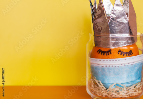 An orange with painted eyes wearing a protective mask against coronavirus and wearing a crown stands in a transparent jar on a bright yellow-orange background with a place for the inscription