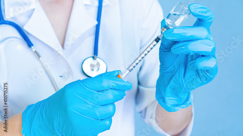 A doctor with a stethoscope in medical gloves is filling a syringe from a vial with medicine or vaccine against coronavirus. Preparation for insulin injection close-up on a blue background.