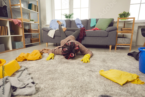 Extremely tired woman lying face down on rug in living-room amid chaos of scattered untidy clothes. Miserable exhausted housewife fallen on floor while cleaning and tidying crazy mess in her house photo