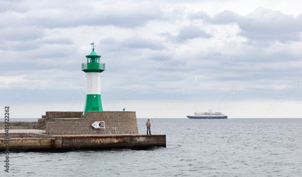 A fisherman stands at the end of the pier in the port of Sassnitz with a fishing rod in his hand. Next to him is the green lighthouse. In the background a ferry drives out to the Baltic Sea.