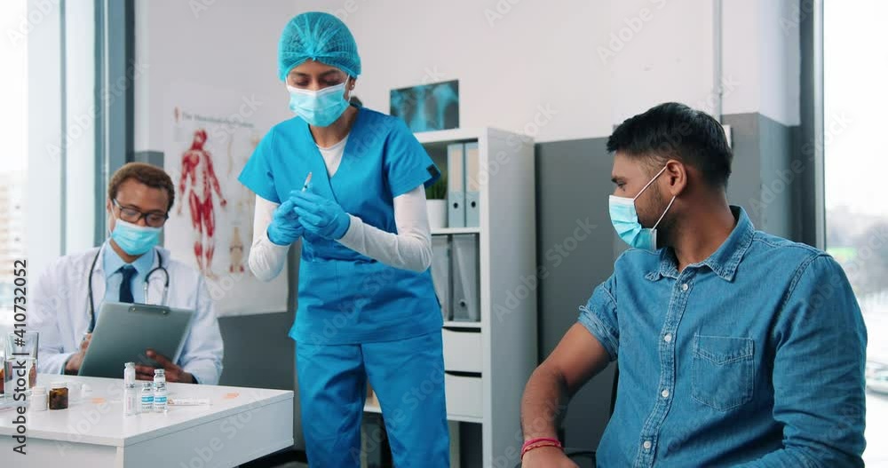 Hindu pretty young female nurse or virologist assistant injecting handsome male patient sitting in hospital cabinet in medical mask while doctor working and reading analysis, coronavirus vaccination Hindu pretty young female nurse or virologist assistant injecting handsome male patient sitting in hospital cabinet in medical mask while doctor working and reading analysis, coronavirus vaccination