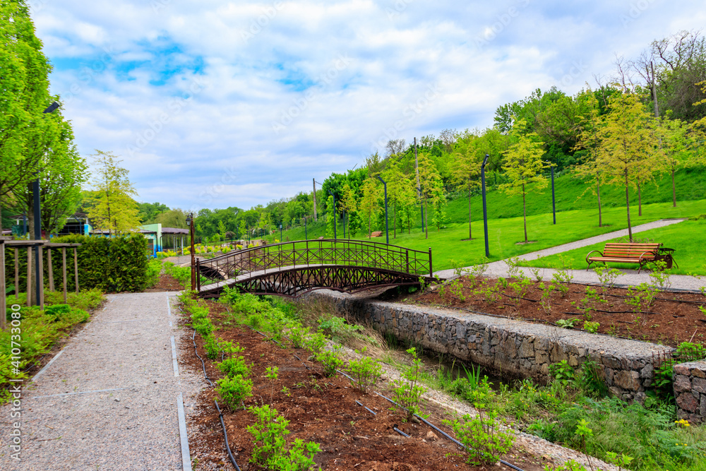 Beautiful view of the city park with arched footbridge across small river and young trees Beautiful view of the city park with arched footbridge across small river and young trees