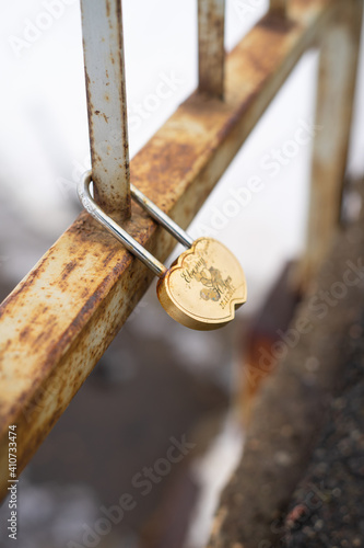 Weathered, heart shaped, golden wedding padlock with name engravings hanging on bridge railing. 