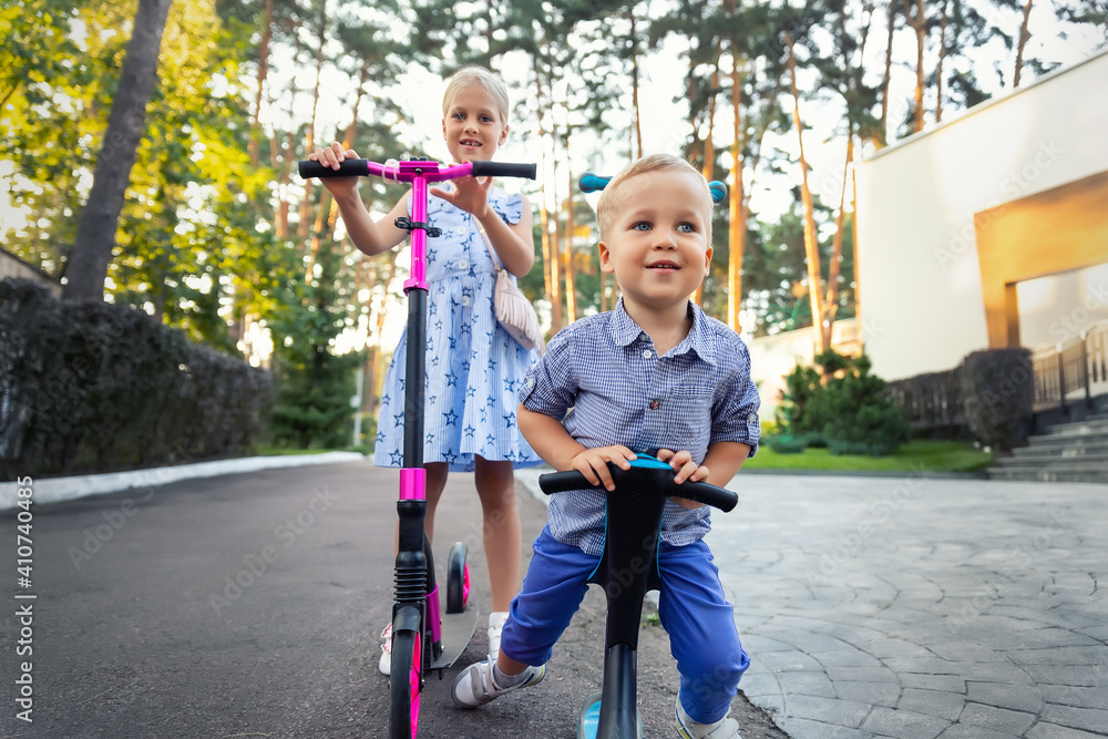 Φωτογραφία Two Cute adorable little caucasian siblings boy in shirt and girl in dress having fun riding three-wheel balance run bike scooter in city park forest