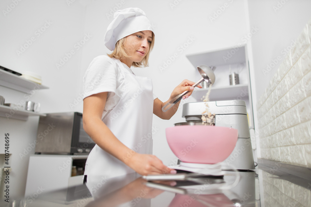 Close up bottom view of young attractive chef woman, adding the flour into pink bowl on electronic weight to make a dough for cake or cookies in her own kitchen or confectionery Close up bottom view of young attractive chef woman, adding the flour into pink bowl on electronic weight to make a dough for cake or cookies in her own kitchen or confectionery
