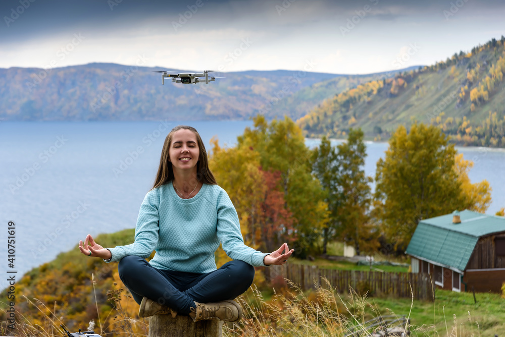 Beautiful young woman having fun with a mini drone outdoors in Lotus pose. The girl sits against the autumn forest background