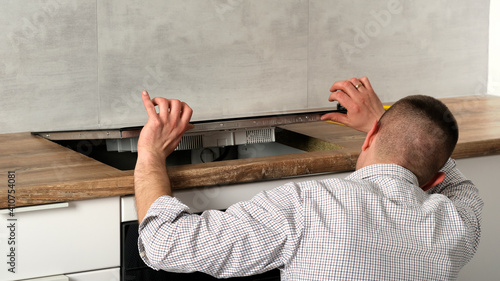A young repairman installs a black induction hob in a modern white Scandinavian style kitchen with a concrete wall. Electrician man, do it yourself. Household chores