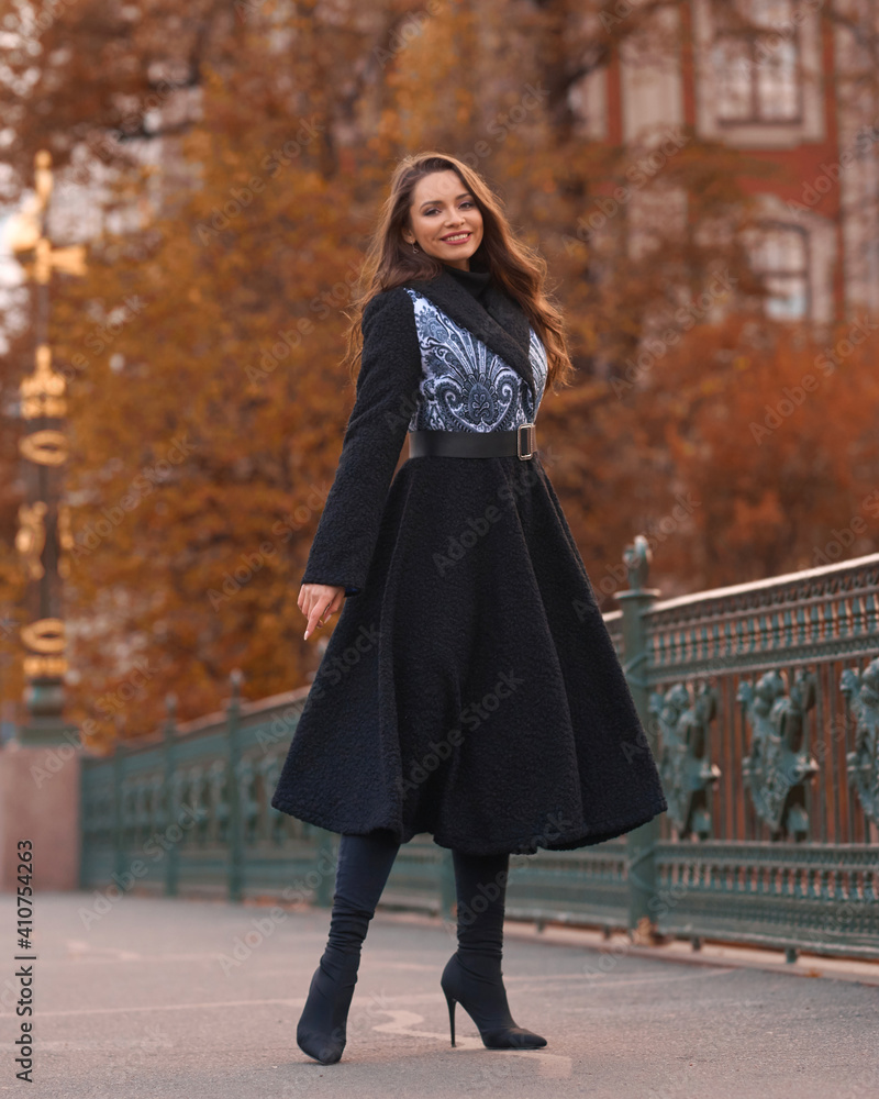 Elegant woman wearing black coat and walking city street on autumn or fall day against town park fence. Pretty girl with makeup and wavy brunette hair