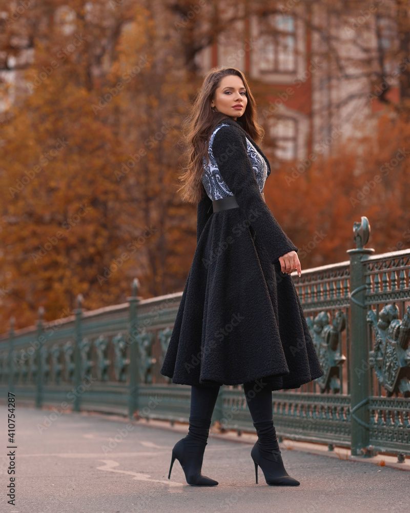 Fototapeta premium Elegant woman wearing black coat and walking city street on autumn or fall day against town park fence. Pretty girl with makeup and wavy brunette hair