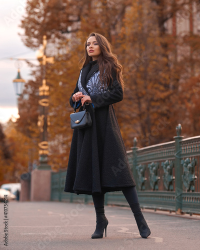 Elegant woman wearing black coat and walking city street on autumn or fall day against town park fence. Pretty girl with makeup and wavy brunette hair