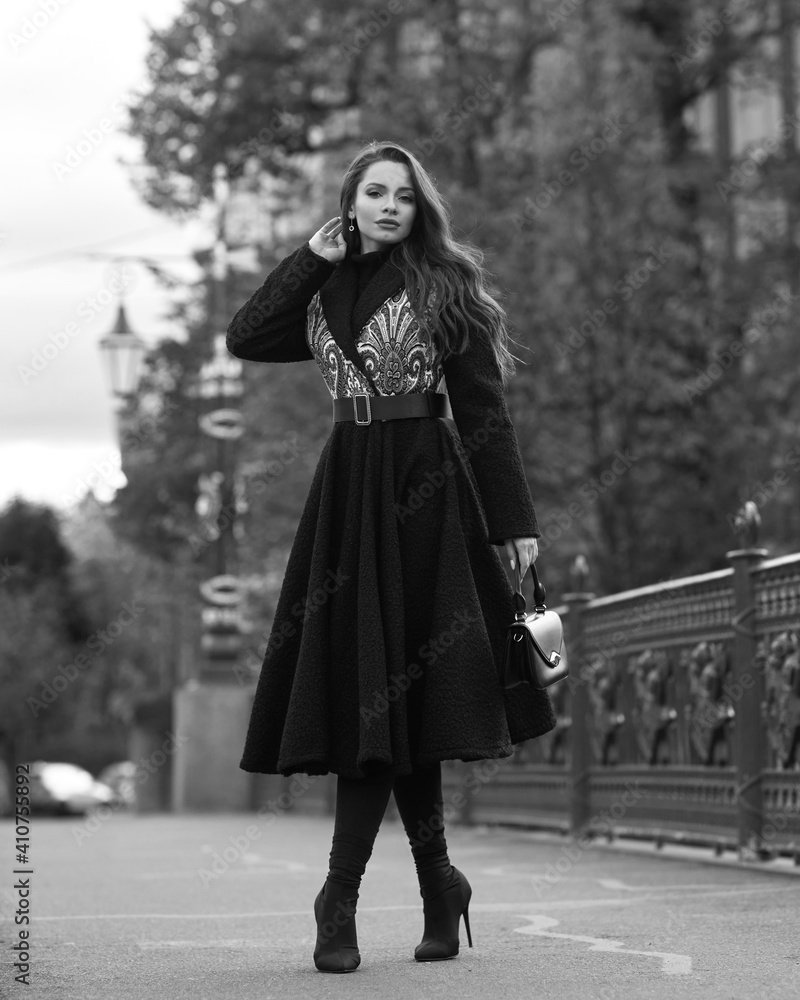 Obraz premium Elegant woman wearing black coat and walking city street on autumn or fall day against town park fence. Pretty girl with makeup and wavy brunette hair