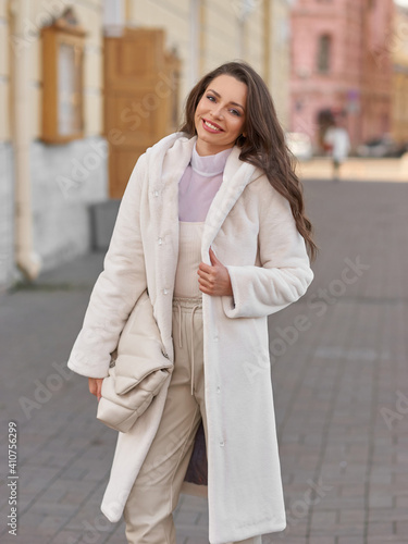 Elegant stylish woman with long brunette wavy hair wearing white trousers, pullover and shoes and white fur coat walking city street on a sunny day