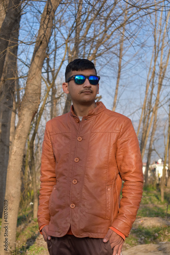 outdoor portrait of indian ethnicity boy looking sideways wearing blue sunglasses and brown leather coat