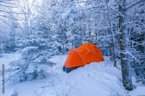 An orange tent stands among the snow-covered trees in the winter forest. Bright tourist tent in the winter taiga