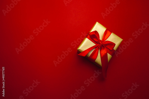 Holiday gift. Box in a gold gift wrapping with a red bow, on a red background. Merry Christmas, Happy New Year and Valentine's Day greetings. Postcard. High quality photo