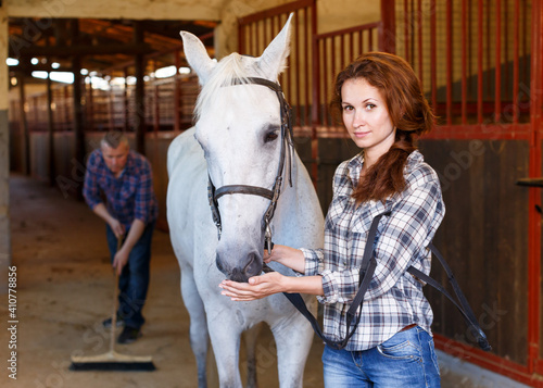 Woman farmer standing with horse, male worker cleaning floor at stabling