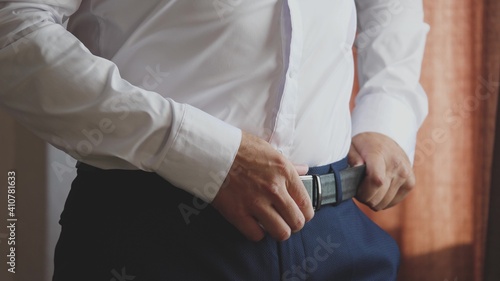 A smart man puts a belt on his pants with a white shirt to keep the pants on his hips. Getting ready early in the morning for work in a room at home. Close-up