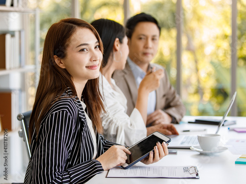 young attractive Asian businesswoman dressed formally sitting and holding tablet with smiley face during working in the office with colleagues sitting in the background