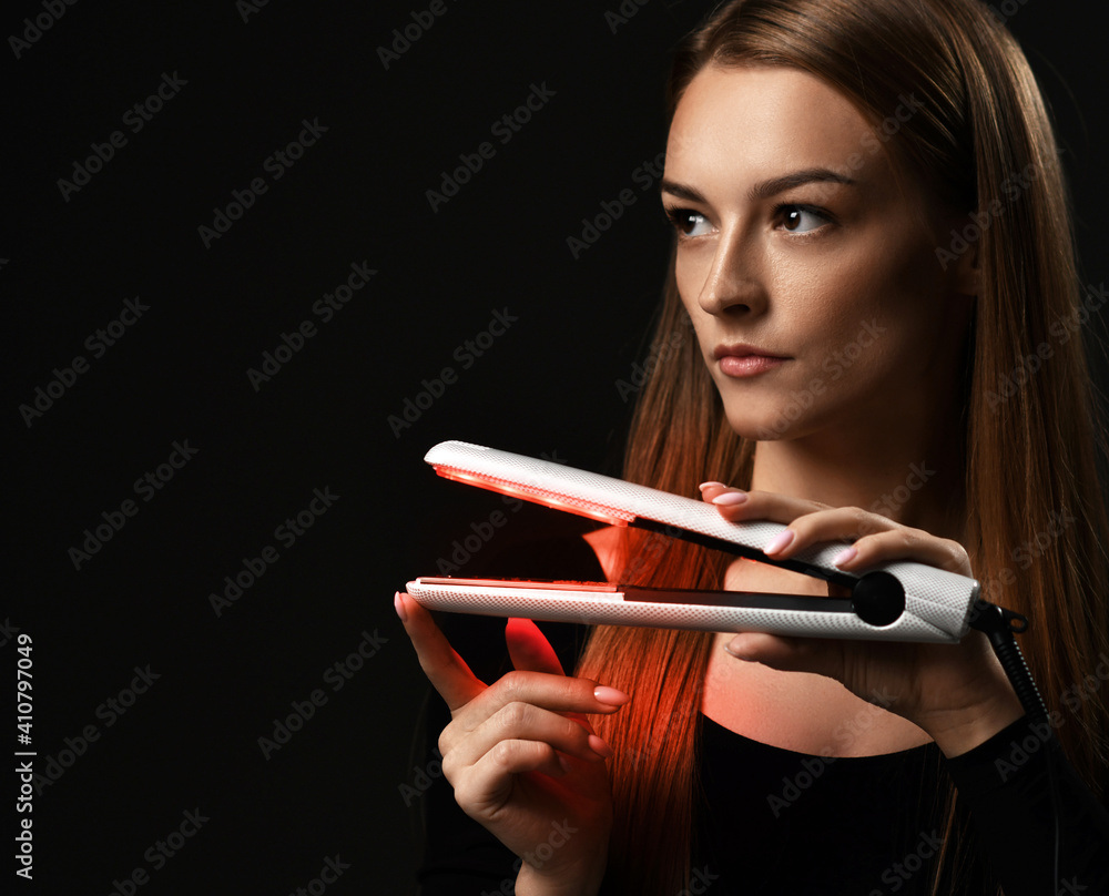 Fototapeta premium Portrait of young refined woman holding modern hair straightener in hands and looking aside at copy space over black background. Hairstyle, hairsalon, hairdresser, fashion concept
