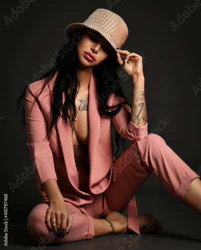 Sexy brunette woman with tattoo on body arms and legs wearing pink silky pantsuit on naked body and hat sits on floor over dark background. Stylish look, fashion, glamour concept photo