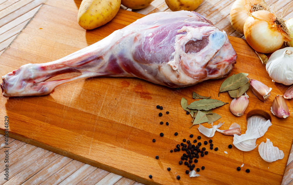 Raw lamb leg on wooden cutting board, potatoes and condiments, ingredients for cooking