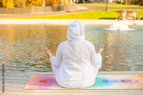 A girl plus a size in a white suit with a hood sits on the shore of the pond and meditates. View from the back. Yoga and wellness concept Copy space